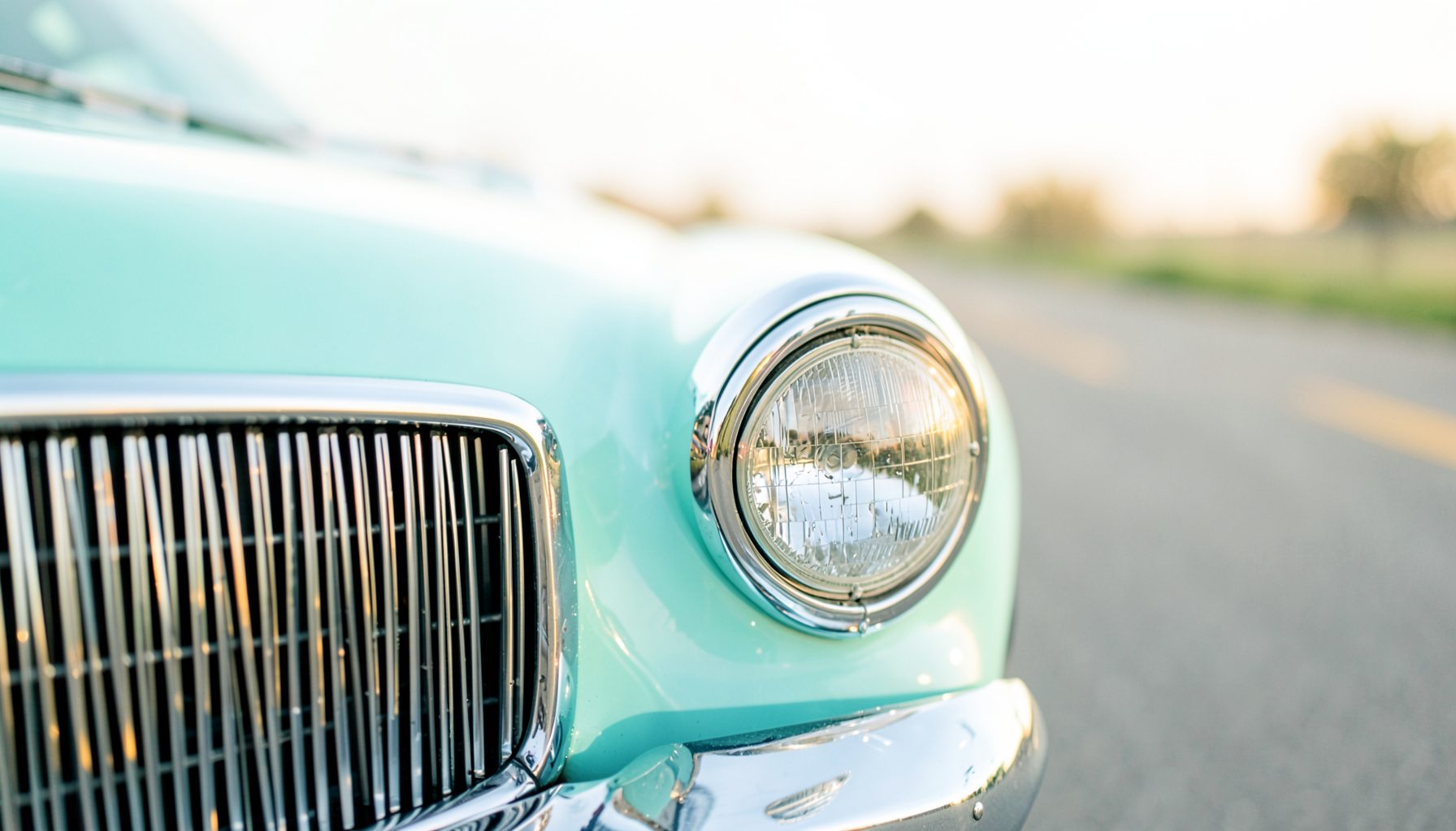 A classic car's polished chrome grille and headlight gleaming under warm evening light, with a softly blurred road stretching into the distance