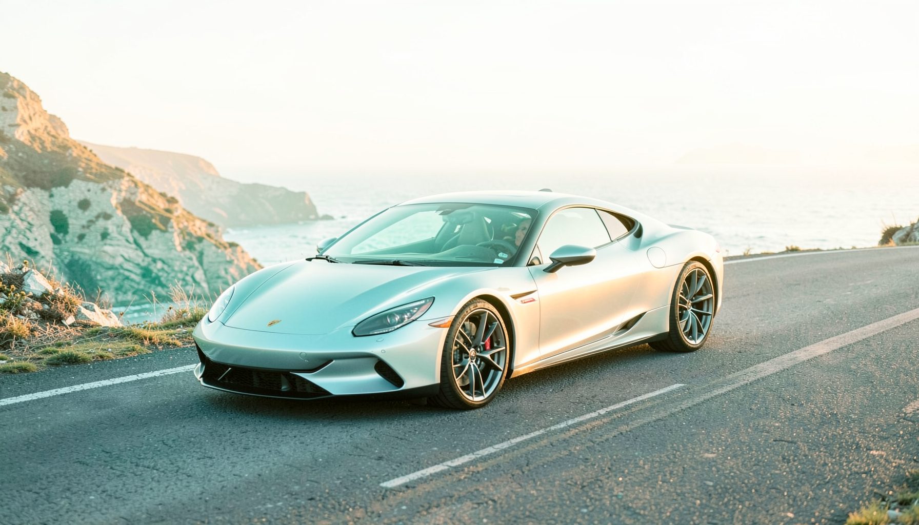 A sleek modern sports car in metallic silver parked on a winding coastal road at golden hour, ocean and cliffs in the background, warm amber light reflecting off the bodywork