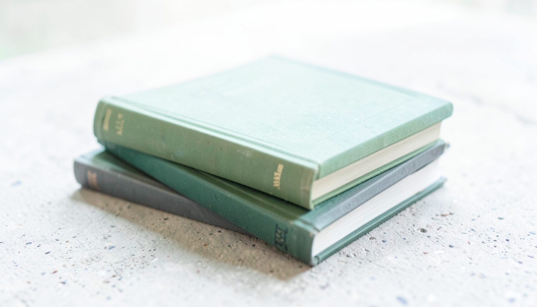A stack of three automotive history books with muted olive and charcoal covers, resting on a concrete surface in soft daylight