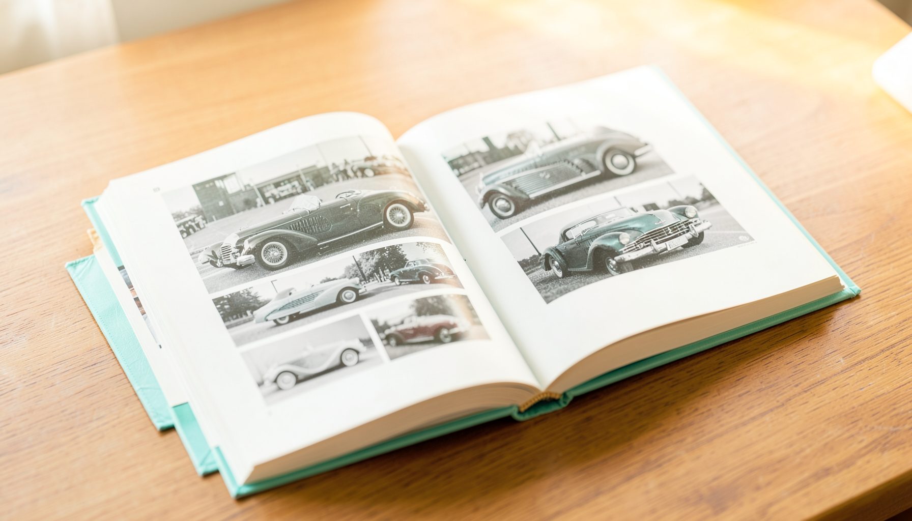 An open hardcover book on a wooden desk, its pages showing black-and-white photographs of classic automobiles, with warm amber lighting